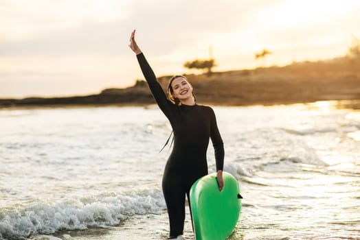 A woman wearing a black wetsuit smiles and holds a green surfboard in the ocean. The water is shallow and the sun is setting in the background, casting a warm glow on the scene. photo