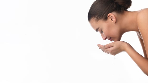 A woman is standing in front of a sink, splashing water on her face with her hands. She is wearing a white t-shirt and has her hair pulled back. The water droplets can be seen on her skin. photo