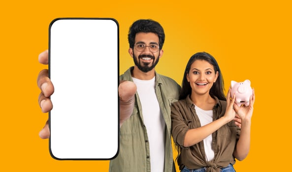 A young Hindu man and woman smile and look at the camera. The man holds a smartphone with a blank white screen and points his finger at the screen. The woman holds a pink piggy bank in her hand. photo
