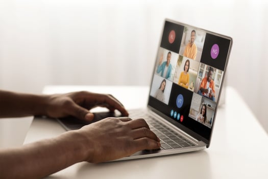 A person is sitting at a desk, using a laptop to participate in a conference with several other people. The laptop screen displays a grid of windows, each showing a different participant photo