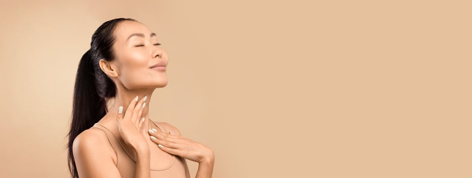 Middle aged Asian woman with closed eyes gently touching her neck, expressing satisfaction with her skin's condition, studio shot with a beige background, copy space photo
