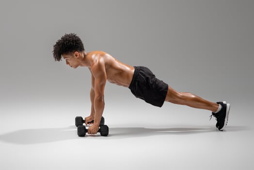 A fit young man engages in a rigorous workout by executing dumbbell push-ups in a well-lit studio gym. He maintains proper form while focusing on strength training and building muscle endurance photo