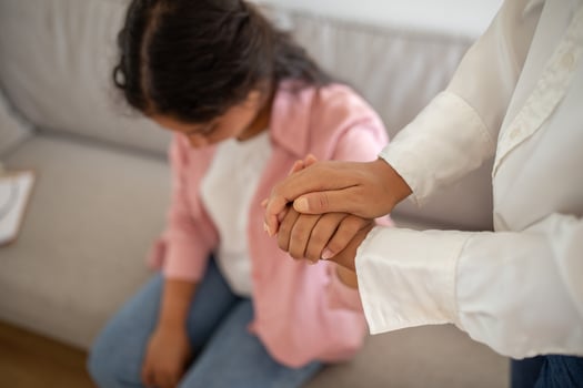 A woman in a white shirt holds the hand of another woman sitting on a sofa, offering comfort and support. The woman receiving comfort has her head down, suggesting sadness or distress. photo