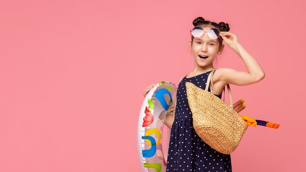 A young girl with her hair in two buns holds a colorful swim ring and a straw bag, ready for a fun-filled summer day, copy space photo