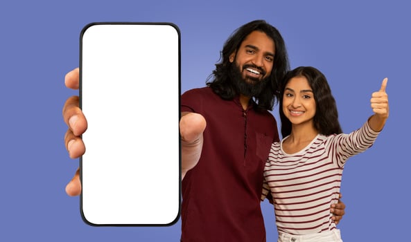 A happy Indian couple poses for a photo, the man holding a smartphone with a blank screen, and the woman smiling and giving a thumbs up. photo