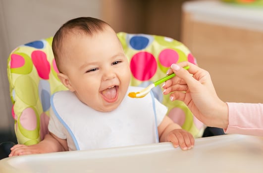 A joyful baby boy sits in a colorful high chair, eagerly opening their mouth as a caregiver feeds them with a spoon of baby food in a bright, cheerful kitchen. photo