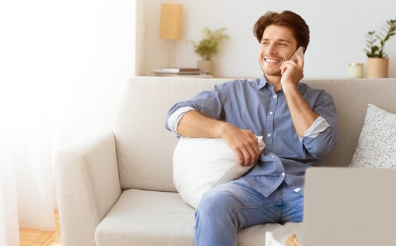 A man in a blue shirt sits on a white couch, smiling as he talks on his phone. photo