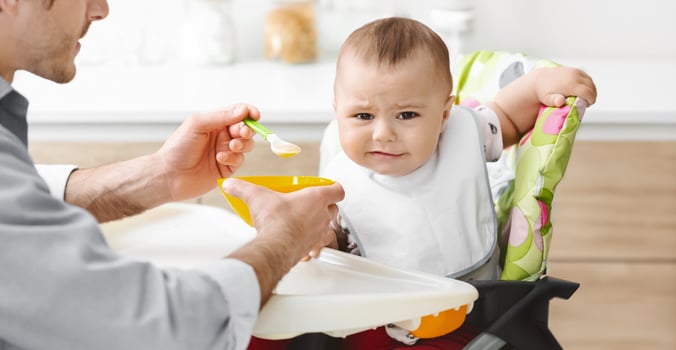 A father is feeding his baby puree from a yellow bowl with a green spoon. The baby is sitting in a high chair with a patterned cushion and is looking at the food with a slightly unhappy expression photo
