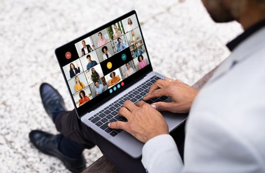African American man sits on a bench with his laptop, engaging in an early morning virtual meeting. Several participants are visible on the screen in a conference format. photo