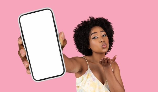 A young black woman with curly hair is holding a smartphone with a blank white screen in her outstretched hand. She is looking directly at the camera and blowing a kiss. photo