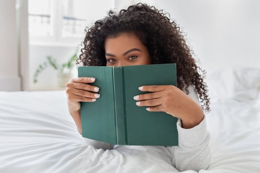 A young Hispanic woman with curly hair is partially hidden behind a green book as she reads, lying on her stomach on a white bed with a look of intrigue or amusement photo