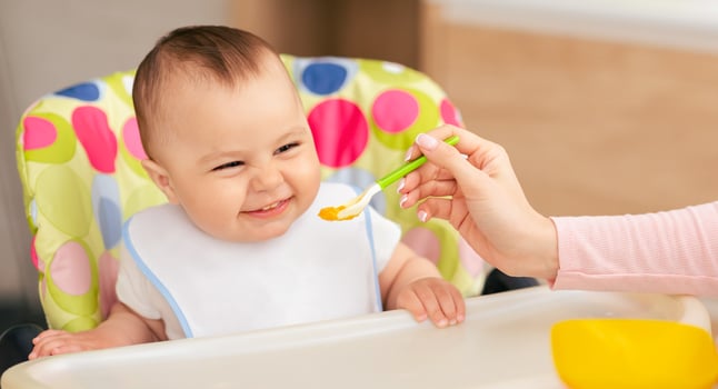 A woman is sitting and feeding a baby with a spoon. The woman hand is holding the spoon, while the baby is looking at the spoon. The baby seems to be enjoying the feeding process. photo