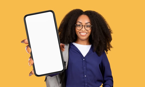 A young black woman with dark hair and wearing glasses holds a smartphone in front of her with a blank screen, smiling and looking directly at the camera. The background is a bright yellow color. photo