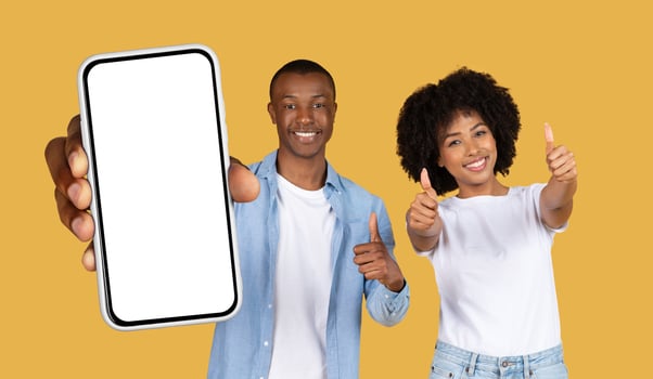 Smiling black couple standing in front of a yellow background. The man is holding a smartphone with a blank screen, while the woman is giving a thumbs-up with both hands photo