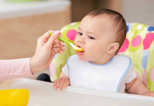 A mother is feeding her baby boy at the kitchen table, with him seated in a high chair. The mood is nurturing and loving, with a homely kitchen setting. photo
