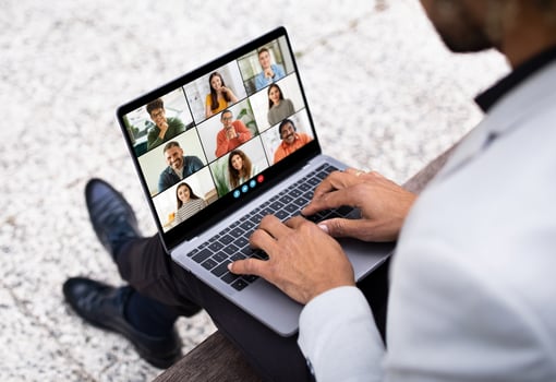 Black man sits on a bench outdoors, using his laptop to participate in a conference meeting. The screen displays a grid of nine participants in various locations, all connected remotely photo