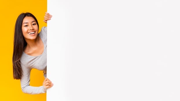 Pretty smiling asian girl looking from behind white advertising board over yellow background photo