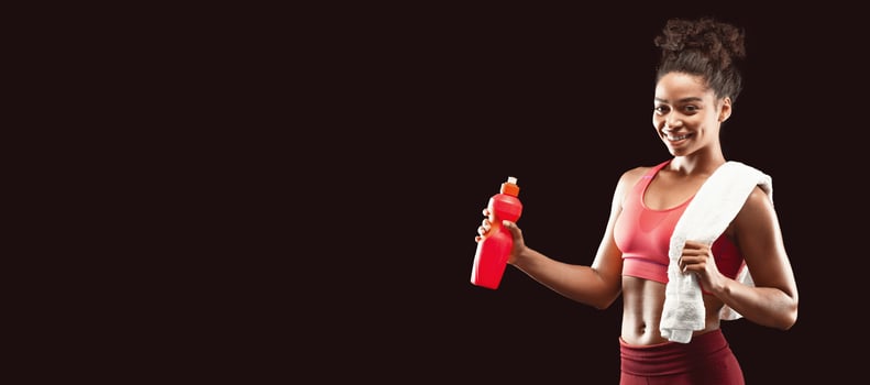Pretty smiling african sporty girl with towel on her shoulders drinking water, black studio background with copy space photo