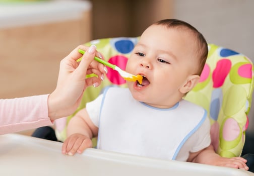 A baby boy is in his high chair enjoying a meal as his mom feeds him. The kitchen setting is comfortable and bright, showcasing a caring and loving interaction. photo