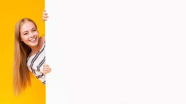 Excited smiling girl looking from behind blank advertising board over yellow background, panorama photo