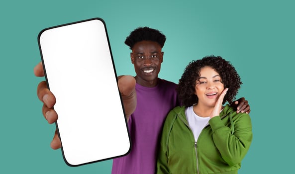 A young black man and woman are standing in front of a teal background. The man is wearing a purple t-shirt and holding a smartphone with a blank screen towards the camera photo