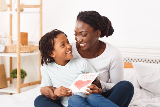Congatulations. Happy african mom embracing her little child and holding greeting card, sitting on bed, copyspace photo