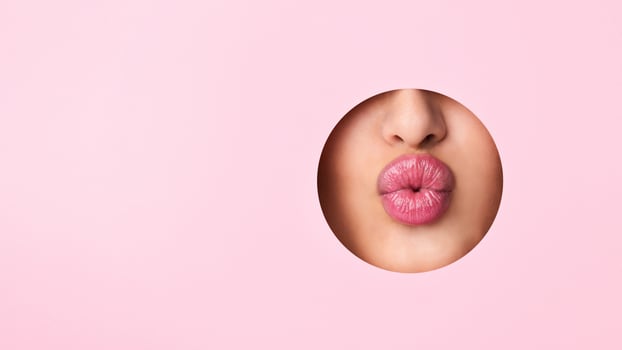 A close-up view of a woman lips protruding out of a hole against a vibrant pink background. The lips appear to be puckered or in a kissing gesture. photo