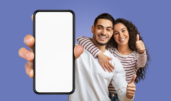 A happy middle eastern couple stands in front of a blue background, the man holding a smartphone with a blank screen toward the camera while the woman wraps her arm around his shoulders photo