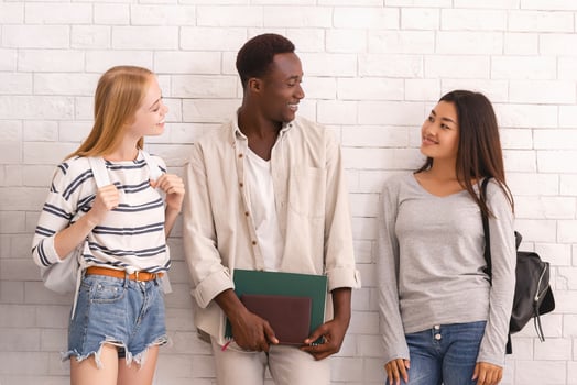 Carefree group of multiethnic students with bags and books having conversation before university over white brick wall photo