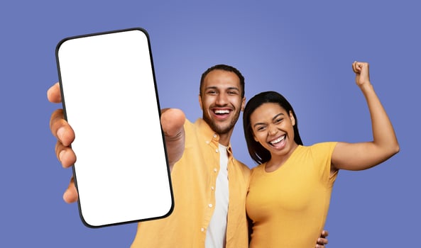 Black man and woman are smiling and looking at the camera while holding a smartphone with a blank screen. The man is holding the phone in front of him, and the woman has her right arm raised photo
