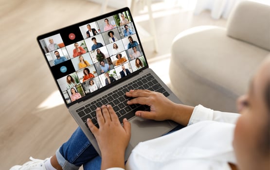 African American lady sits on a couch, holding a laptop in her lap. The laptop screen displays a conference with a grid of numerous participants. photo