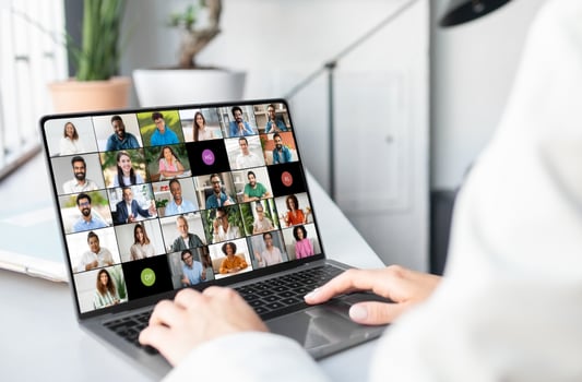 A woman is seated in front of a laptop, engaging with a group of people visible on the screen. She appears focused and attentive, possibly in a virtual meeting or online conference, cropped photo