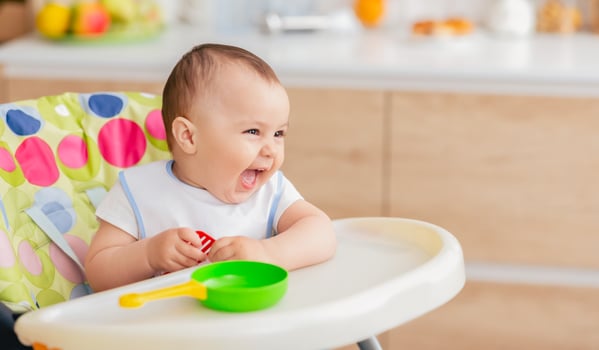 A baby is joyfully laughing while sitting in a high chair with a green bowl and yellow spoon in front of them. The setting appears to be a kitchen, with various kitchen items photo