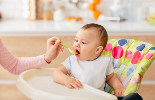 A woman is sitting and feeding a baby with a spoon. The baby is holding onto the spoon and eating from it. The woman is gently guiding the spoon to the baby mouth. photo