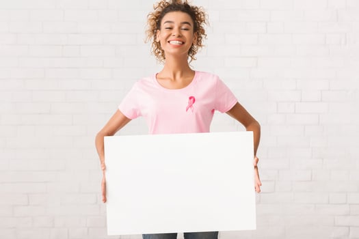 Breast Cancer Treatment. Afro Volunteer Girl In Pink T-Shirt With Awareness Ribbon Holding Blank Poster Standing Near Wall Indoor. Mockup photo