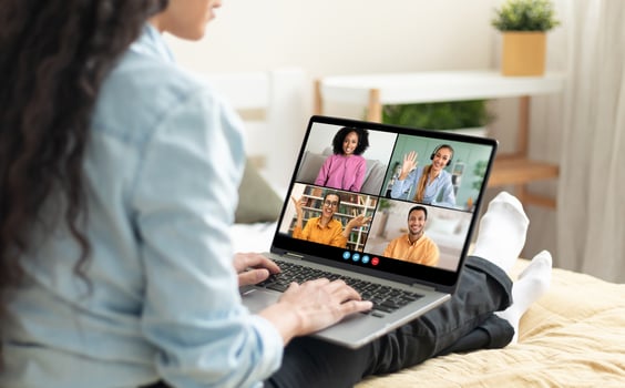 A woman sits on a bed in her home, using a laptop to chat with friends. She is smiling and typing on the keyboard. The laptop screen shows four people in different windows photo