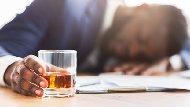 Alcoholism, drinking concept. Drunk african employee sleeping at working place, holding glass with alcohol, selective focus, panorama photo