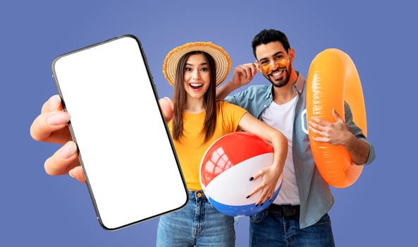 A young arabic couple is excited about their summer vacation. The woman is holding a beach ball and smiling, while the man is holding an inflatable ring. They are both pointing to a smartphone photo