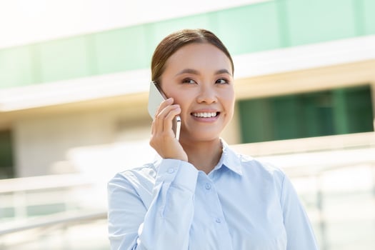 A young Asian woman with a cheerful expression is engaged in a phone conversation while standing outdoors. She is dressed in a light blue shirt and appears to be enjoying her discussion. photo