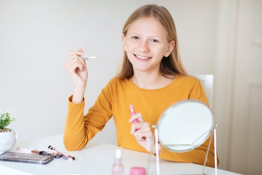 A teen girl with long blonde hair sits at a table and applies lip gloss while looking in a small mirror. Her hands are steady as she uses the lip gloss applicator to apply color to her lips photo