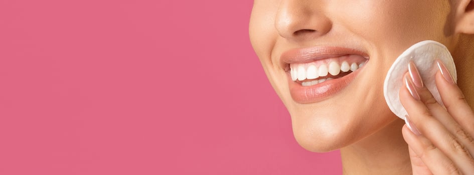 A close-up photo of a smiling woman applying a cotton pad to her cheek. The woman teeth are white and bright, and she has a youthful glow. The background is a vibrant pink, copy space
