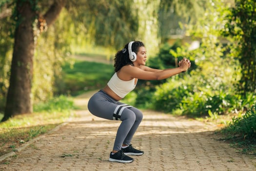 A woman wearing headphones is squatting on a paved path. She appears focused on something, with her hands resting on her knees. The surroundings are nondescript. photo