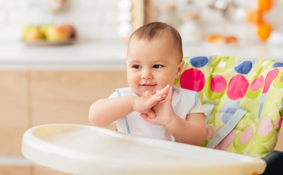 A baby is seated in a high chair in a kitchen, surrounded by kitchen appliances and utensils. The baby is looking around curiously, with a bib around its neck. photo