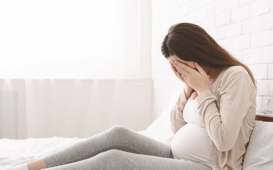 A pregnant woman sitting on a bed with her hands covering her face. She appears to be in a moment of contemplation or distress. photo