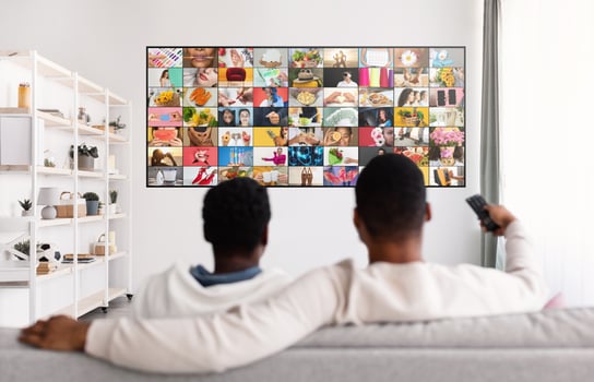 Two men sit on a white couch facing a large flat-screen television with a grid of various images on the screen. One man is holding a remote control in his right hand. photo