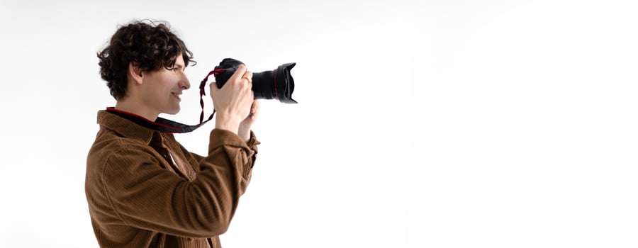 A young man with curly hair is enthusiastically taking pictures using a professional camera in a brightly lit studio. His focus is evident as he aims the lens towards the subject, copy space photo