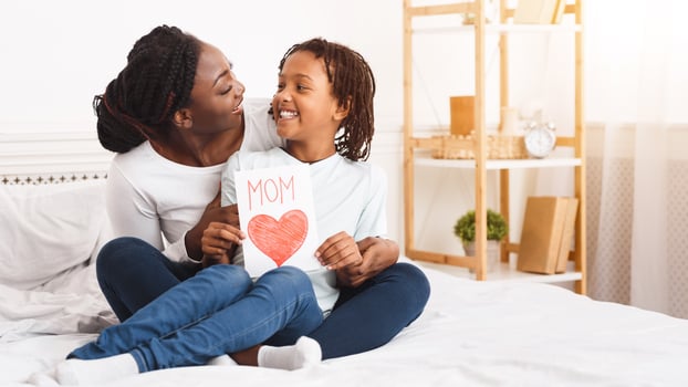 I Love You, Mommy. Happy black mother hugging her little daughter and holding greeting card, sitting on bed, free space photo