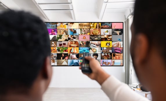 A man is shown from behind, sitting in front of a television displaying a multitude of different channels. He holds a remote control in his hand, presumably changing channels or adjusting the volume photo