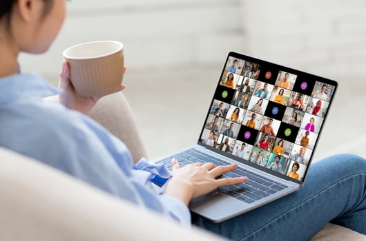 A woman is sitting on a couch, holding a cup of coffee in one hand and using a laptop with a conferencing app open. The screen shows numerous participants in the call. photo