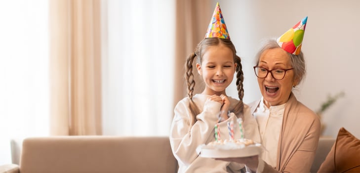 A young girl wearing a party hat proudly holds a birthday cake while her grandmother expresses excitement and joy. The cozy home setting enhances the celebratory atmosphere. photo